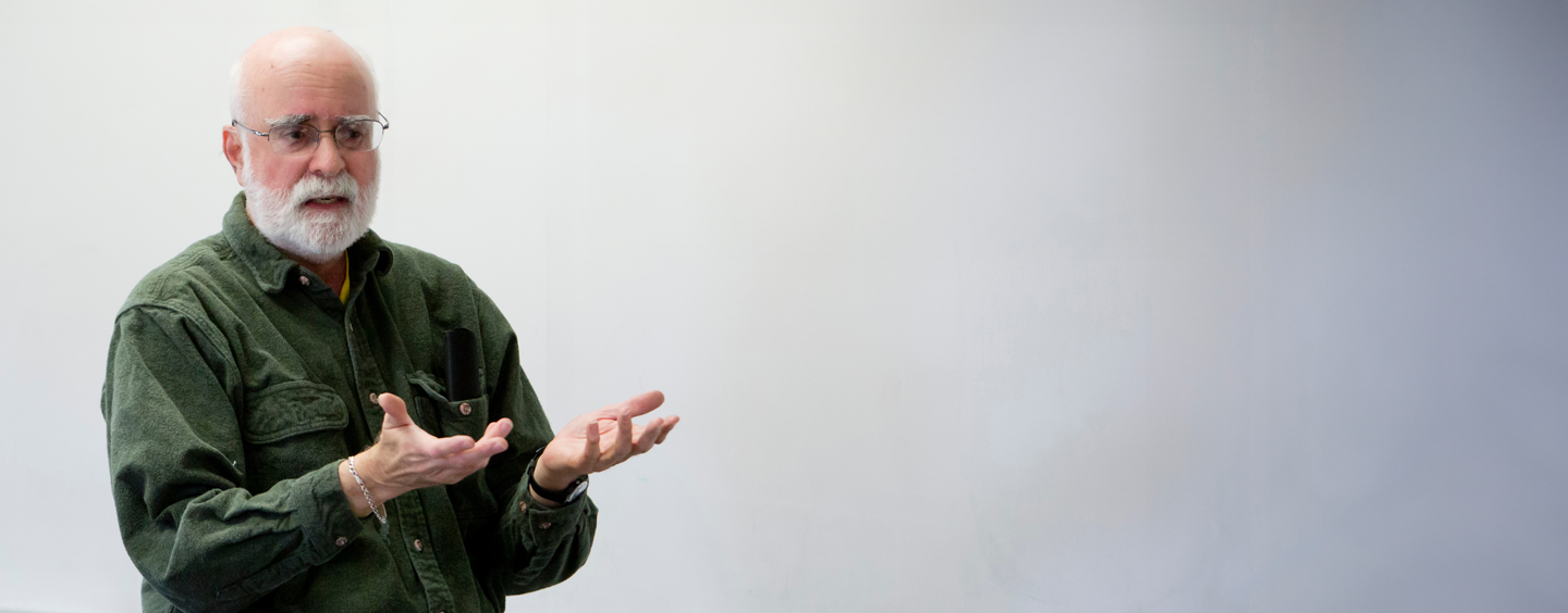 A professor standing at the front of a classroom, from the waist up, in front of a blank whiteboard.